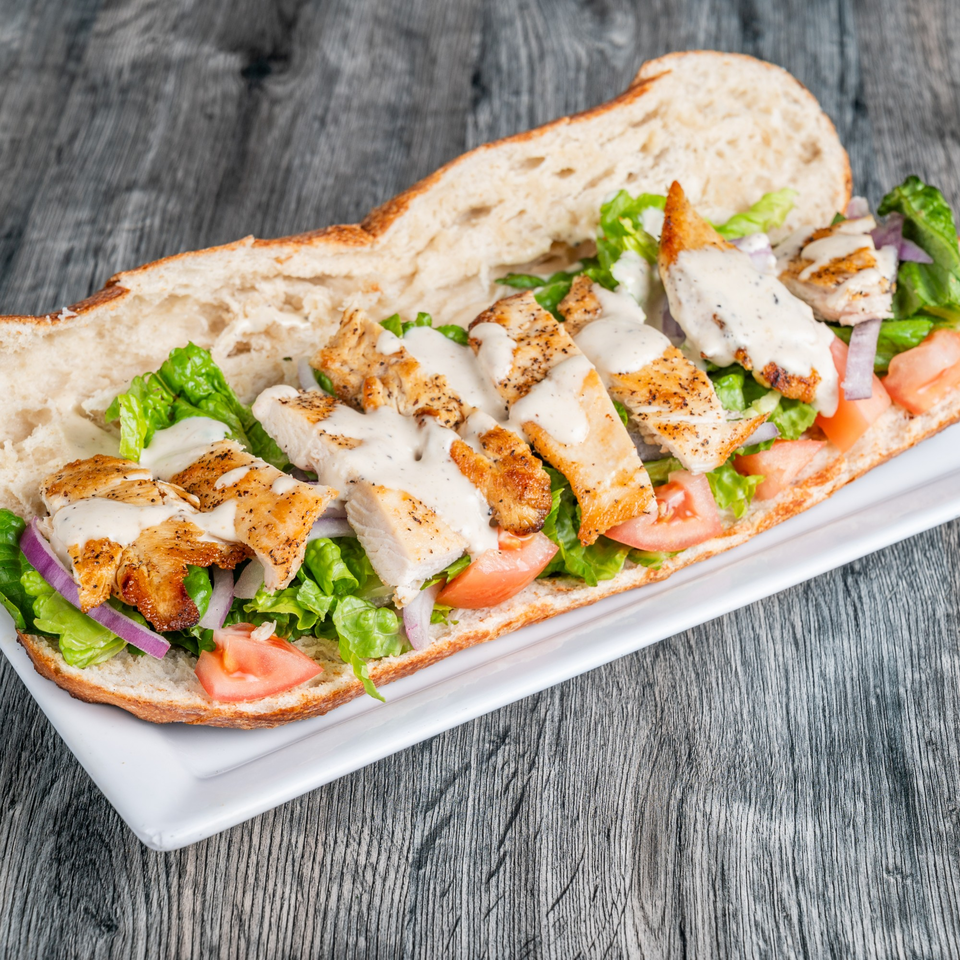 Grilled chicken sub sandwich with lettuce, tomatoes, red onions, and creamy dressing on a white plate.