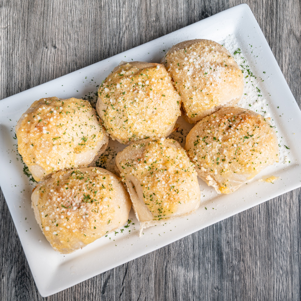 Six golden-brown Fantasy Pizza garlic knots, sprinkled with Parmesan cheese and parsley, on a white rectangular plate.